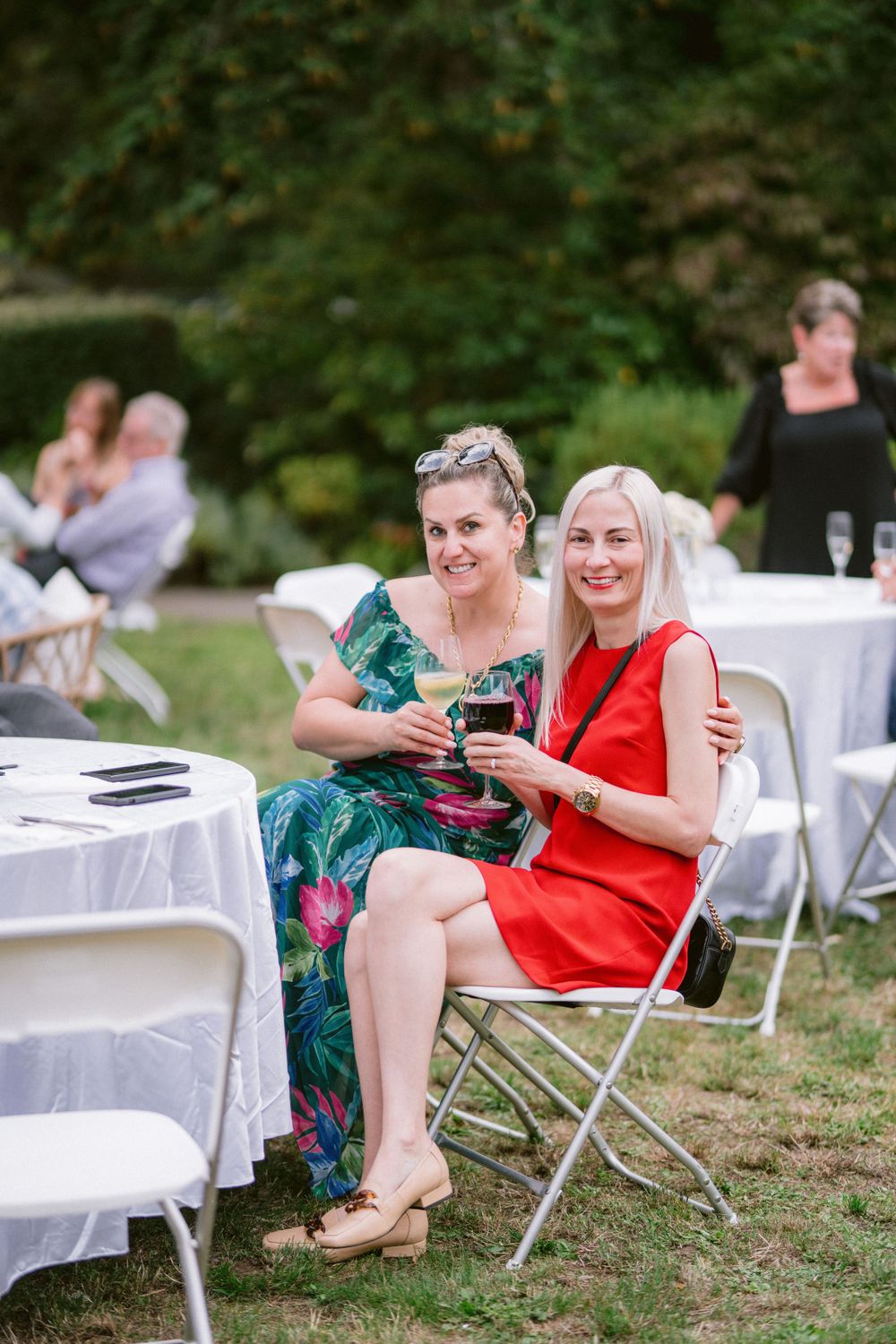 Two women in colorful dresses sit at white tables during an outdoor event.