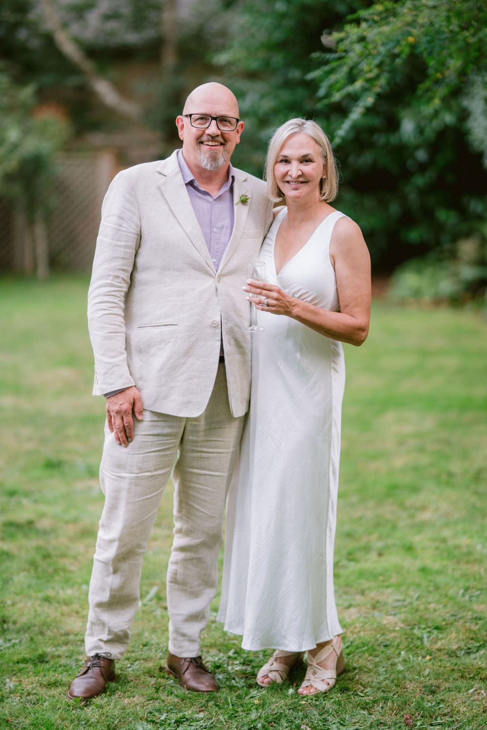 A couple in matching light-colored formal wear pose together on a lawn.