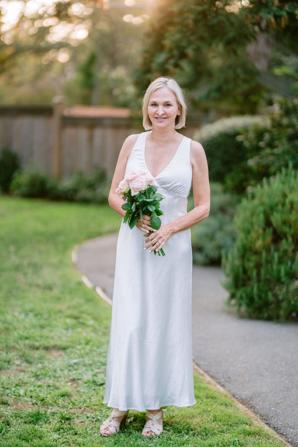 A bride in a flowing white dress holds a small bouquet while standing on a garden path at sunset.