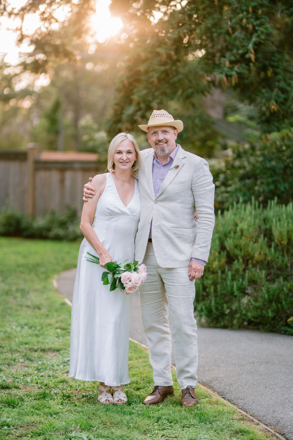 A wedding couple poses together on a garden path in matching light colored attire at golden hour.