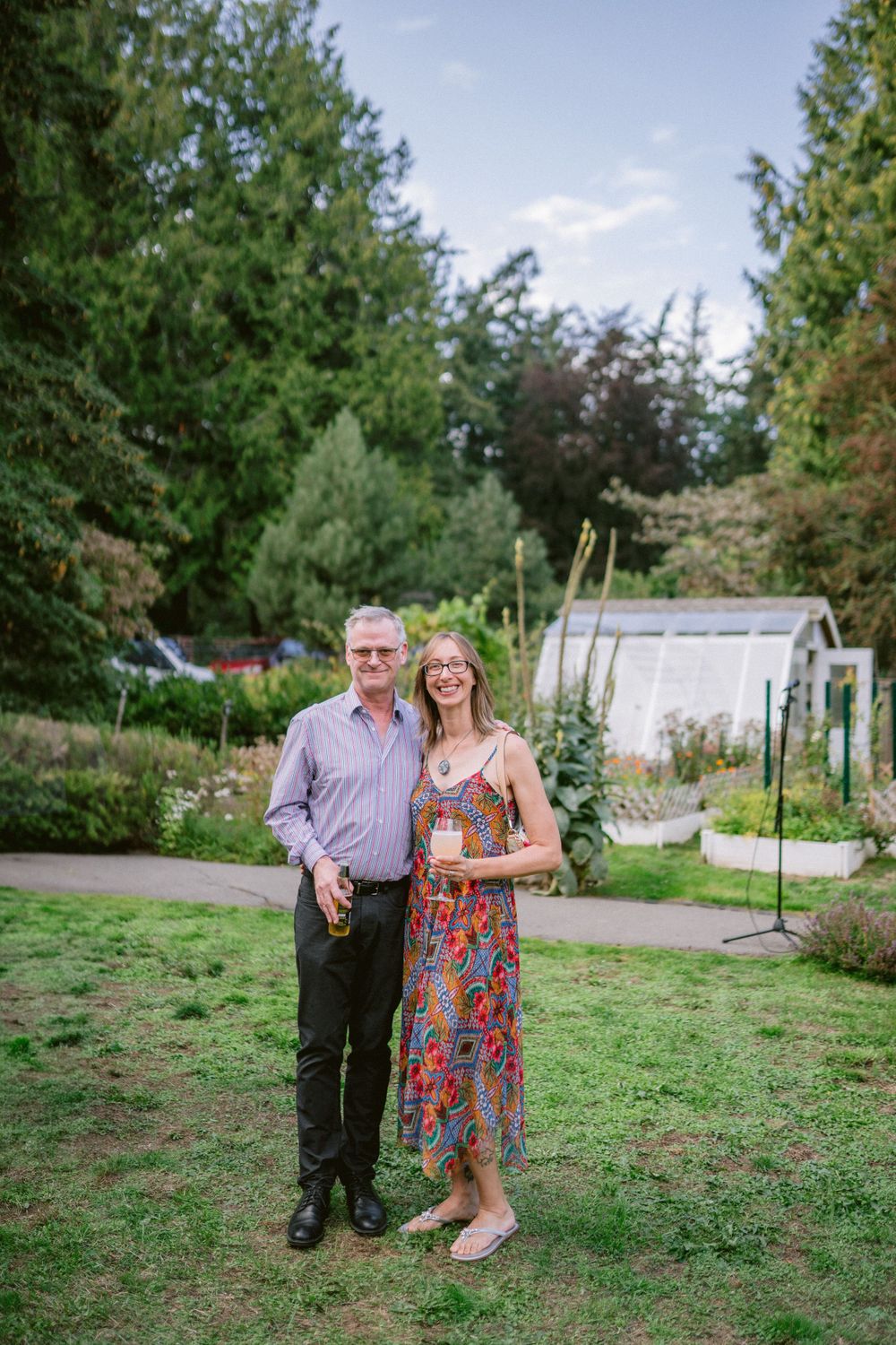 A couple poses together in a lush garden setting with a greenhouse in the background.