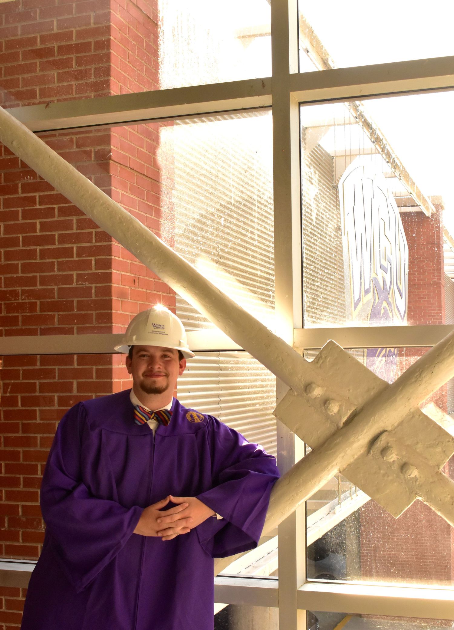 Graduate in purple academic regalia posing by structural beam in sunlit building interior.