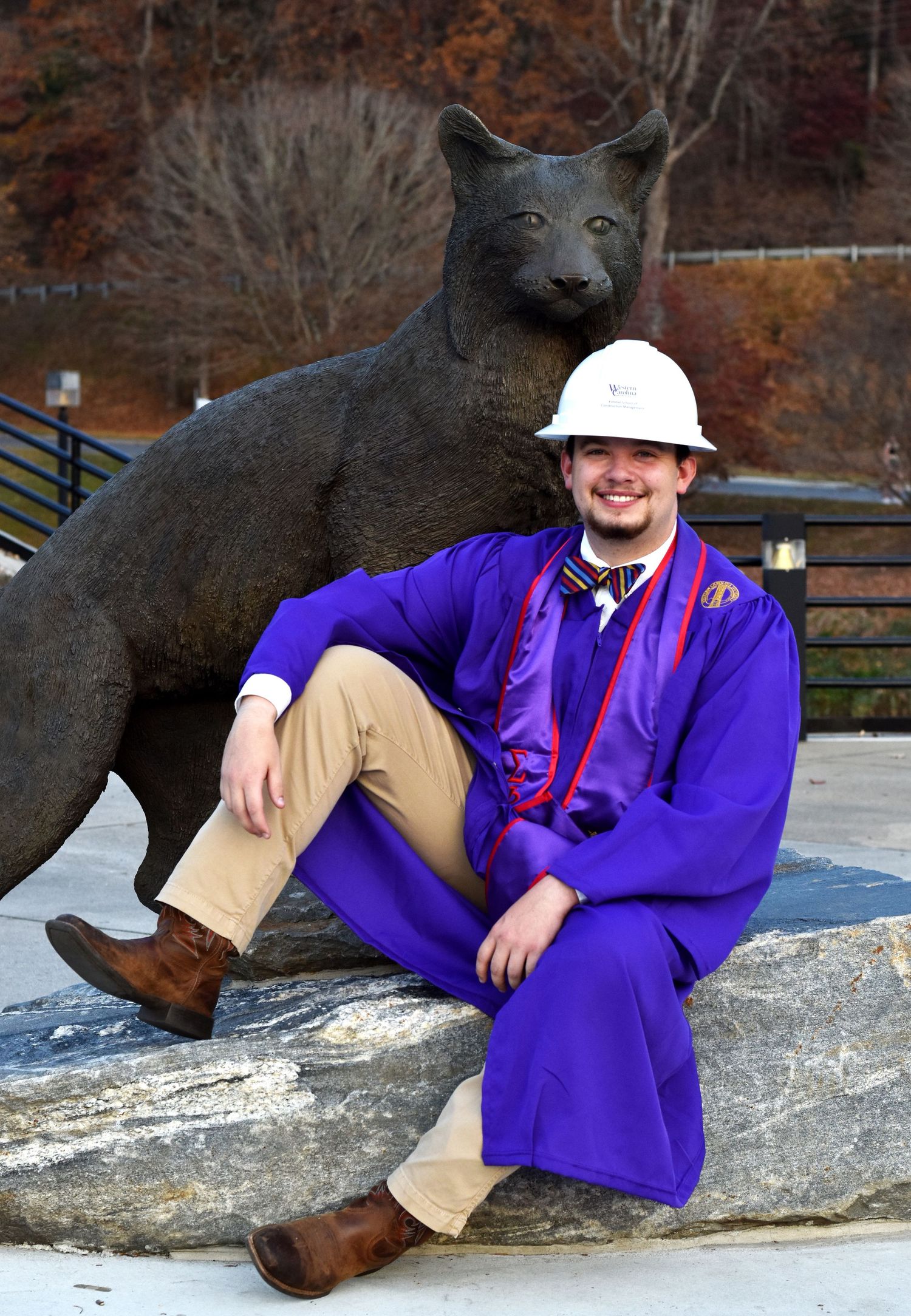 Graduate in purple regalia sitting next to large cat statue on outdoor campus grounds.