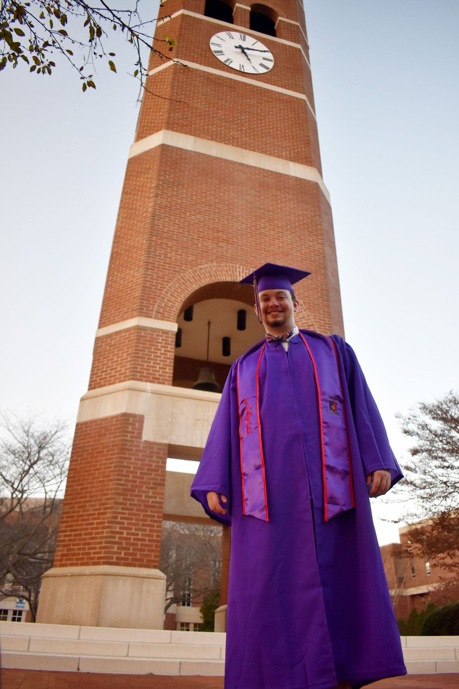 Graduate in purple regalia standing proudly in front of brick clock tower.