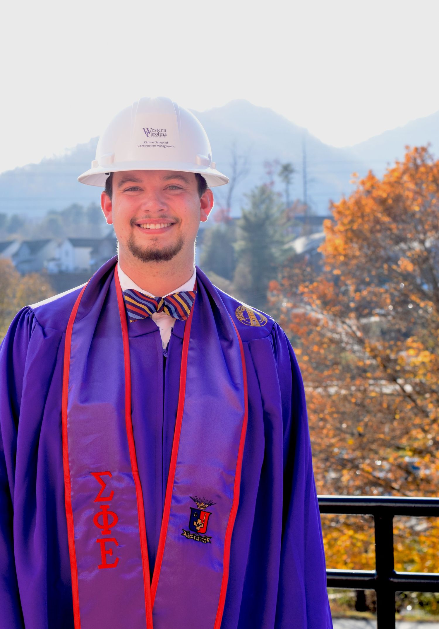 Graduate in purple regalia poses outdoors against autumn mountain backdrop.