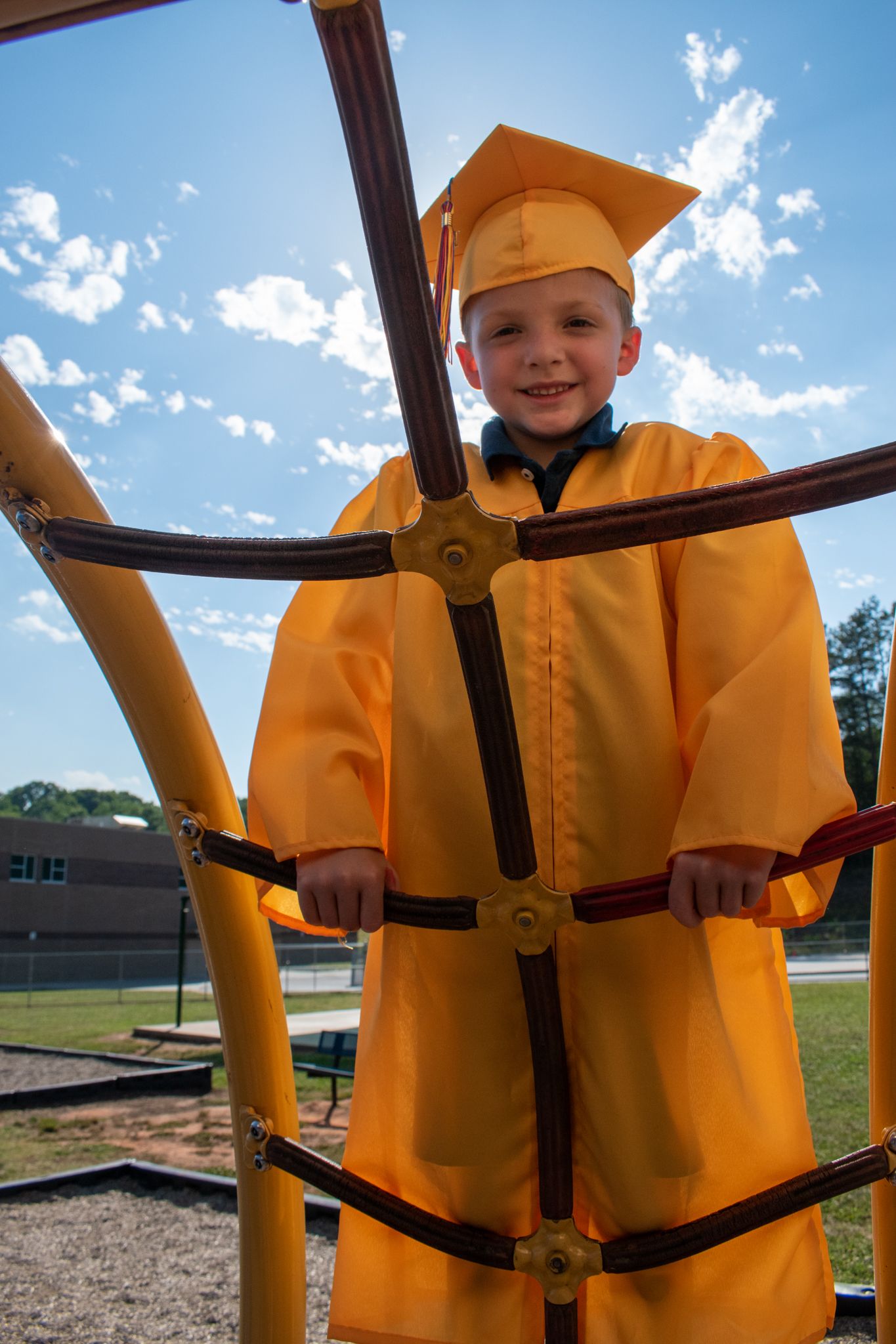 Child in yellow graduation cap and gown climbing on playground equipment.