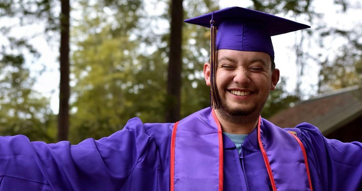 Graduate in purple cap and gown celebrating outdoors with arms outstretched.