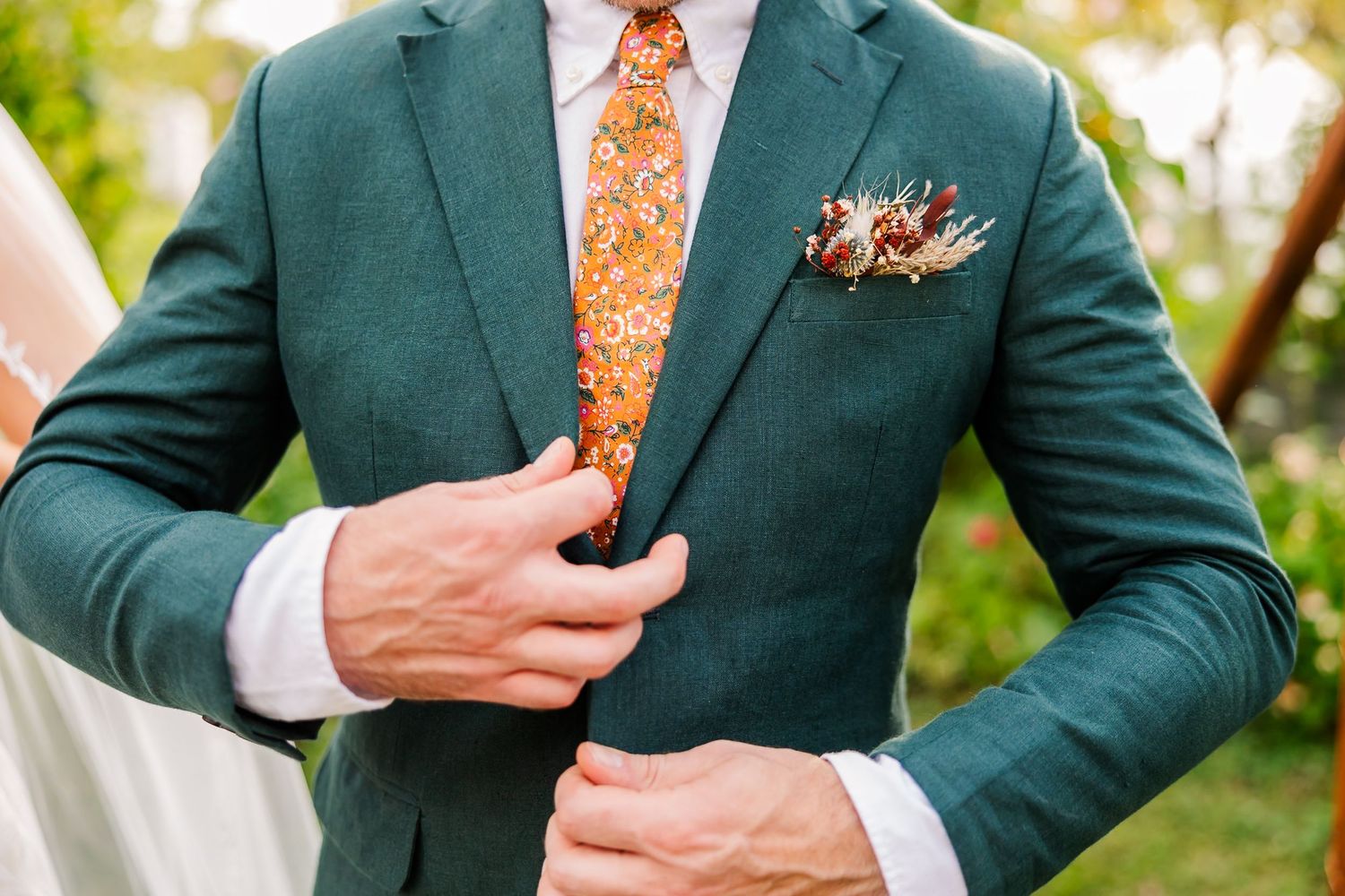 A groom in a dark green suit with orange tie holds a boutonniere during an outdoor wedding ceremony.