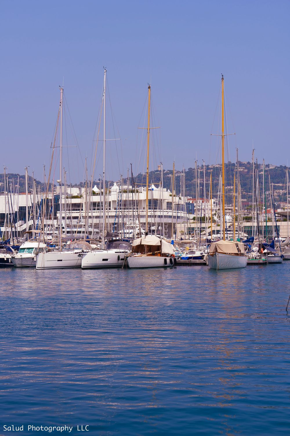 A marina filled with sailboats and yachts on calm blue waters with buildings visible along the shoreline.