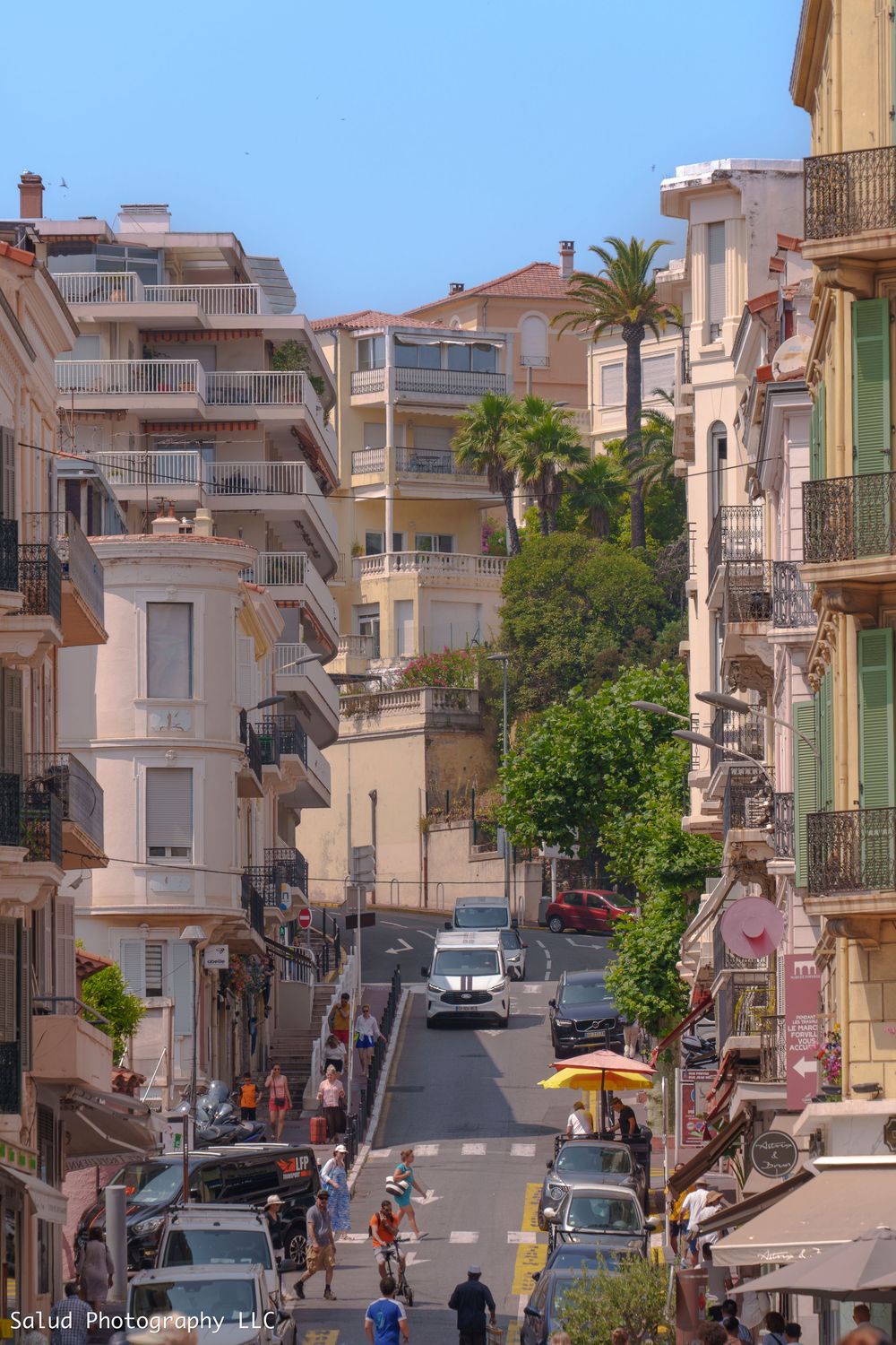 Steep hillside street in a Mediterranean coastal town with white buildings and palm trees lining the road.