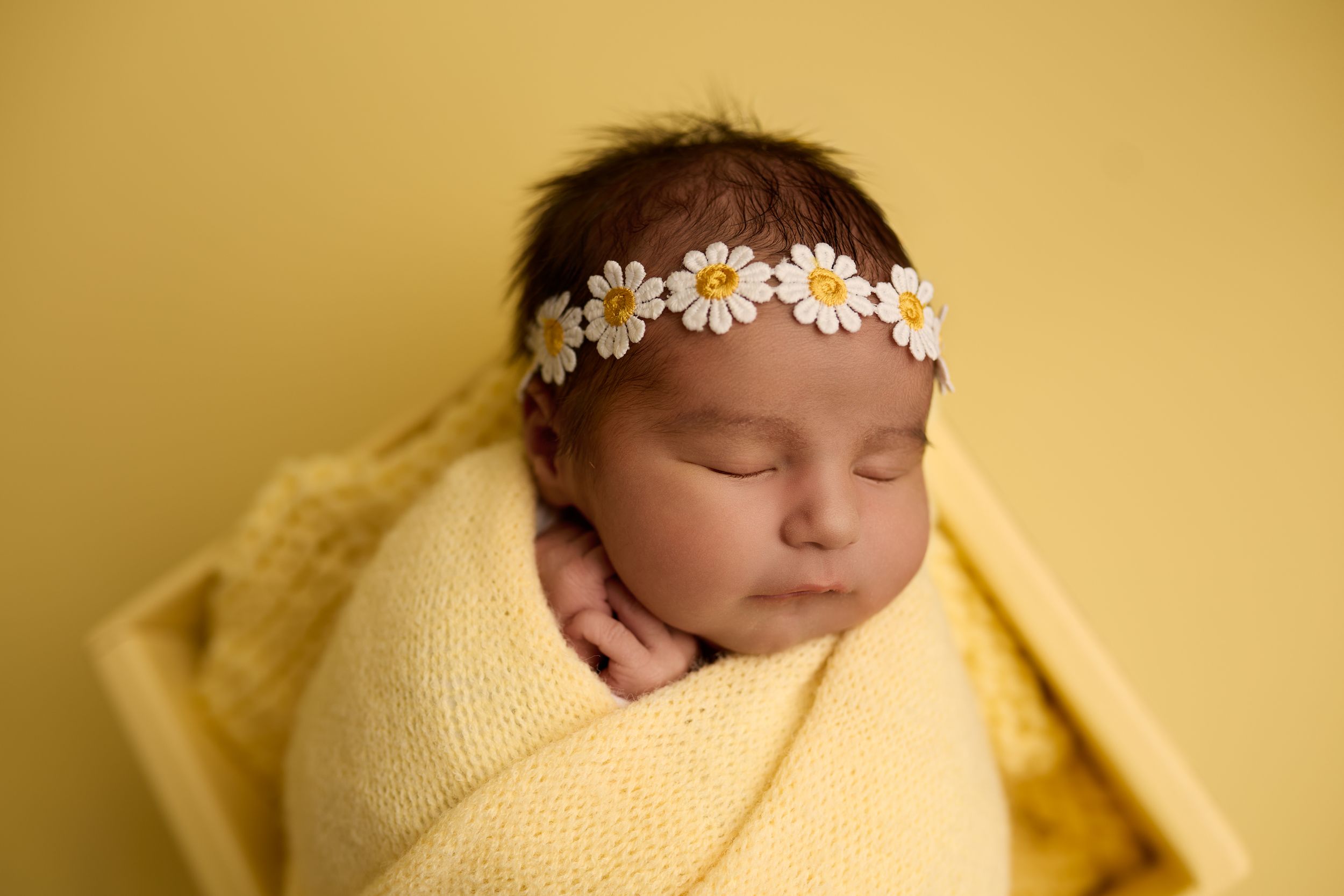 Newborn baby sleeping peacefully wrapped in soft yellow blanket with delicate floral headband.