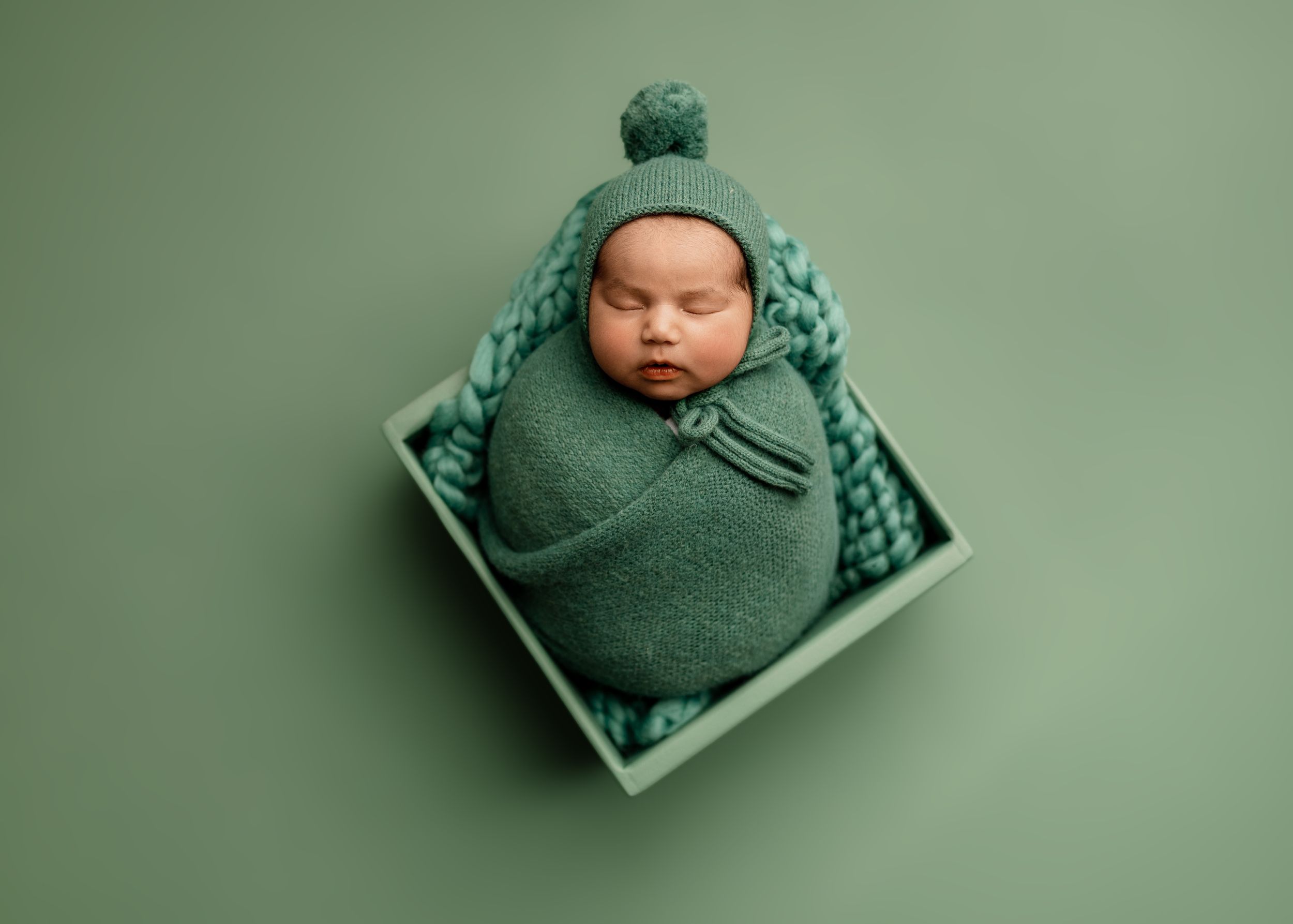 Newborn baby swaddled in green wrap wearing knit hat, sleeping peacefully in a green wooden box on green background.
