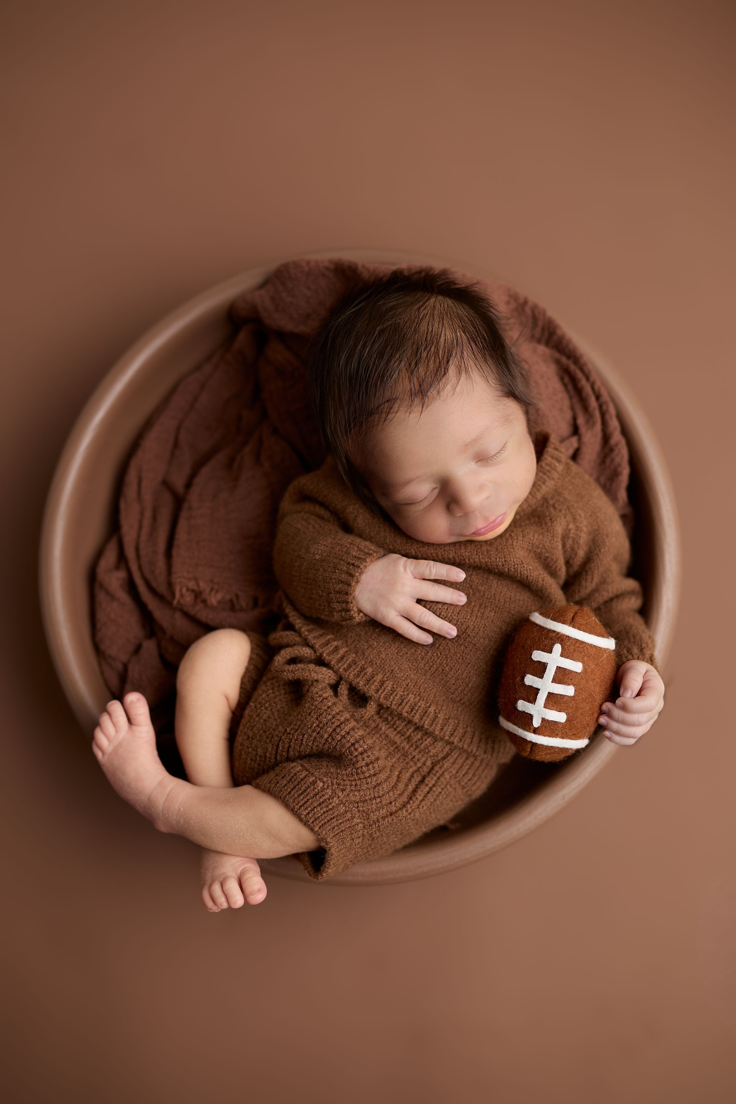A newborn baby swaddled in brown knit fabric with a small football prop creating a sports-themed photography series.