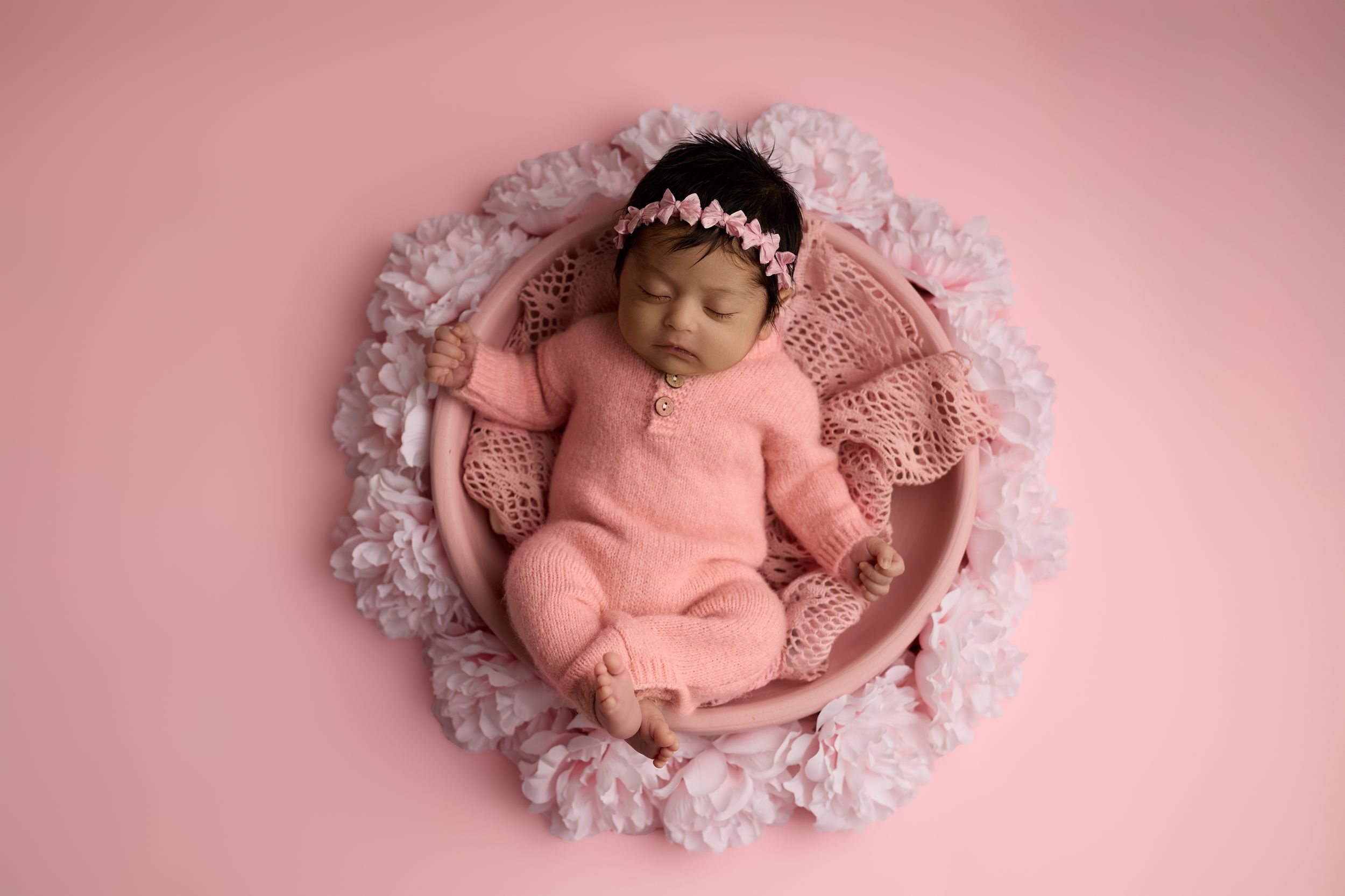 Sleeping newborn baby in pink outfit lies curled up on white lace doily against soft pink background.