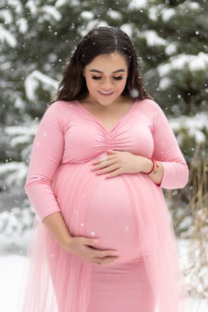 Maternity photo of a girl with dark hair looking down at her stomach while wearing a tight baby pink dress