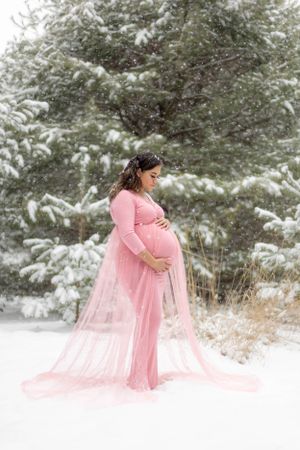 Maternity photo of a dark haired girl holding her belly in the snow wearing a light pink dress