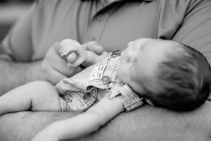Black and white photo of a baby being held in his dads arms holding onto his dads finger while sleeping