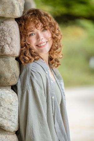 Girl with red curly hair leaning against a stone pillar looking into the camera