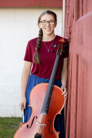 Senior photo of girl with hair in a braid holding her cello leaning against the barn