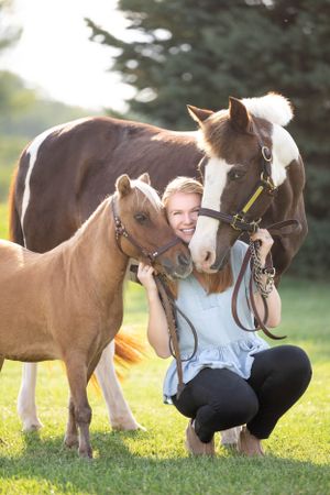 Beautiful strawberry blond girl cuddling with her mini pony and paint pony as she crouches in the grass
