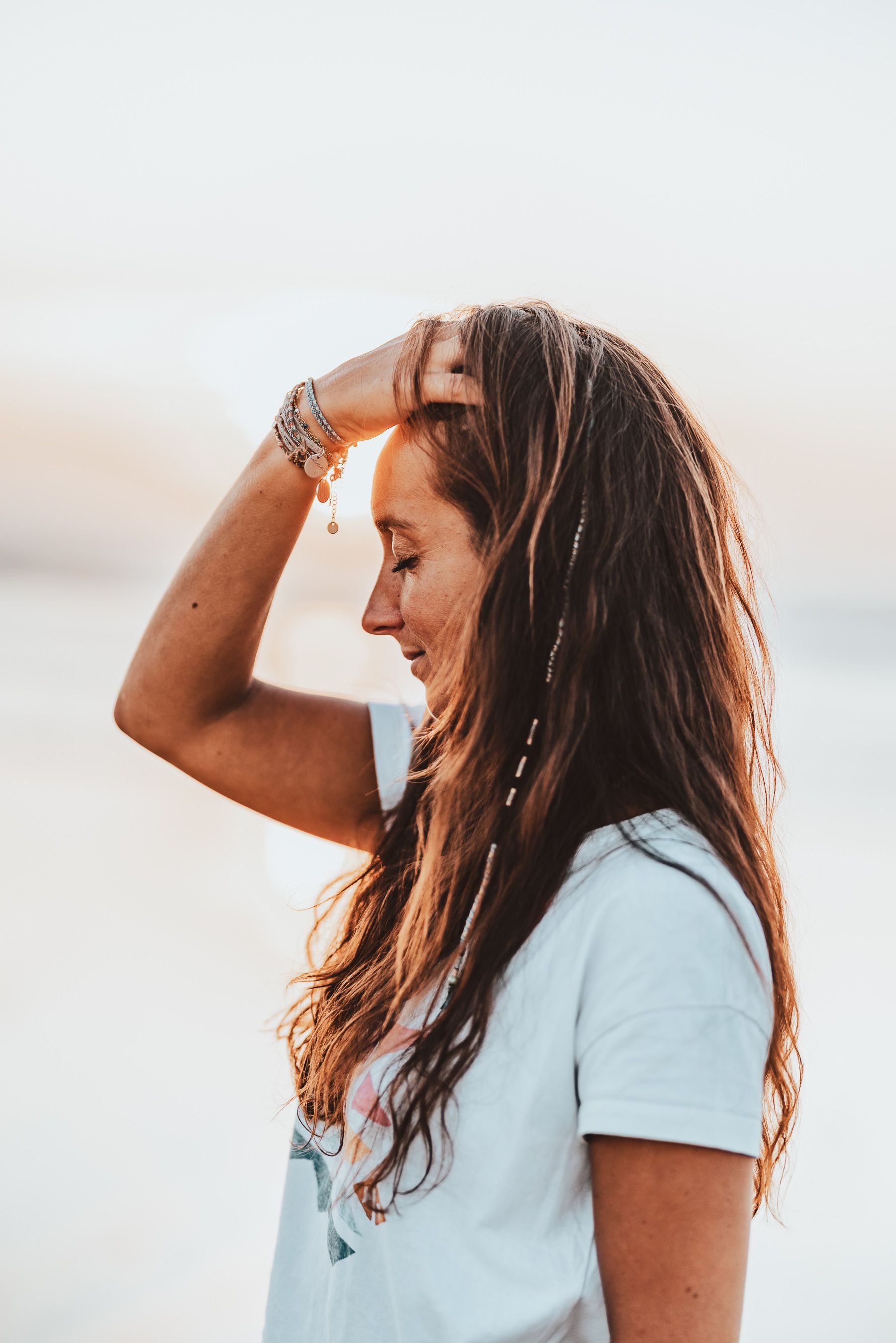 Woman in light blue t-shirt laughs joyfully while running fingers through her long wavy brown hair at sunset.