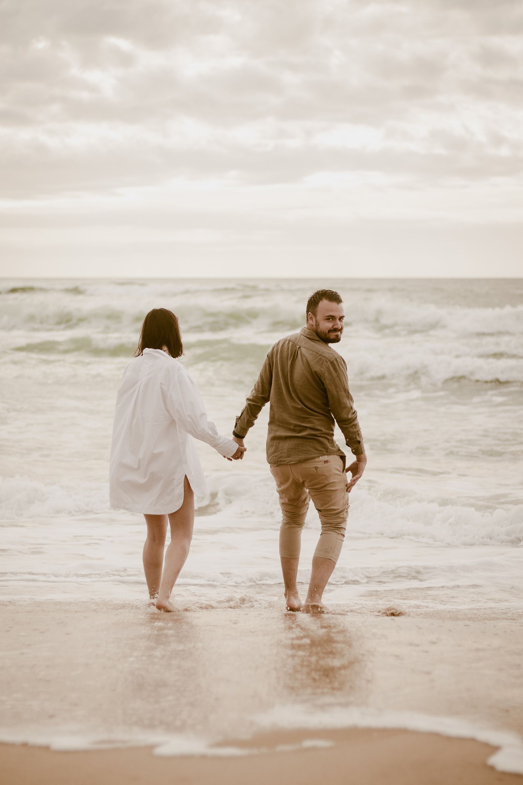 Two people wade in shallow ocean waves at sunset while wearing casual clothing.