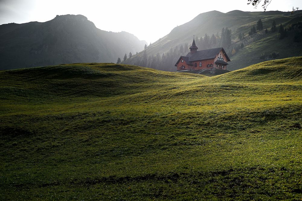 Scenic mountain landscape with rolling green meadows and a rustic cabin nestled against misty alpine peaks at sunset.