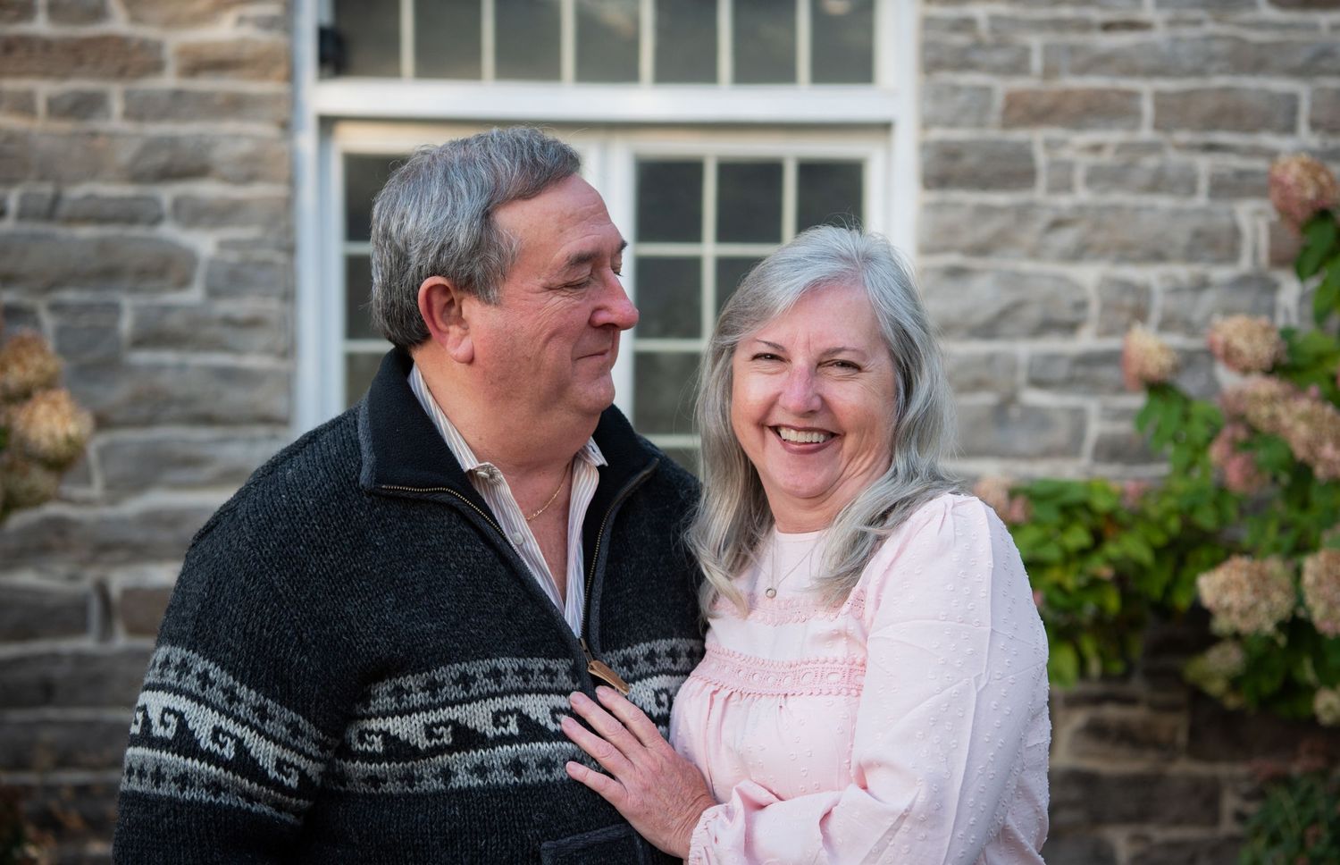An elderly couple in winter sweaters share tender moments across multiple candid outdoor photos against a stone wall.