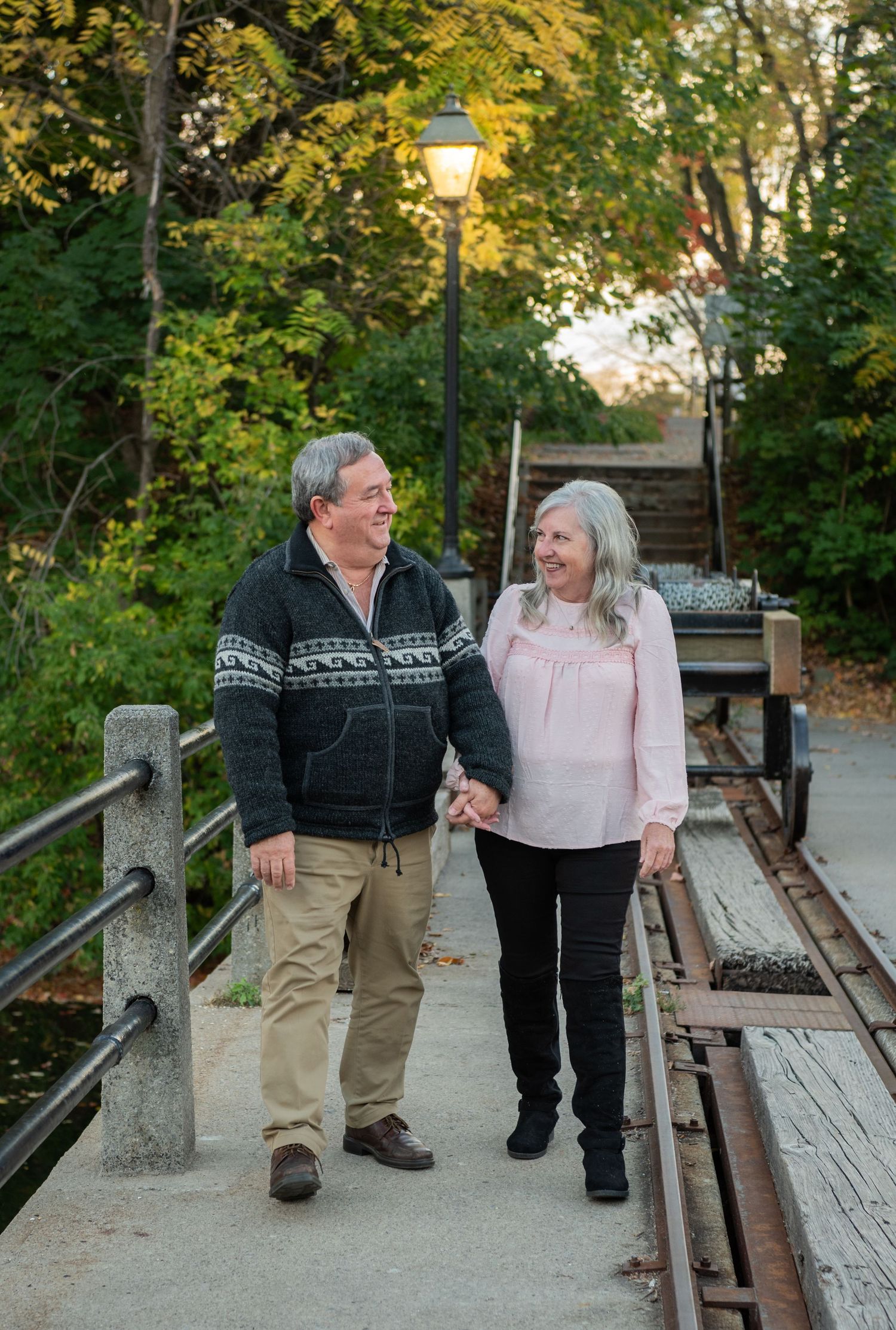 A couple in matching sweaters shares a tender moment while walking along a rustic wooden boardwalk in autumn.