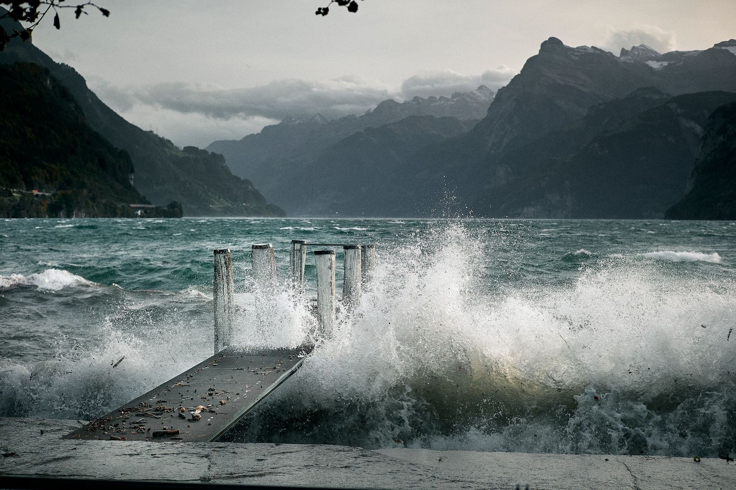 Dramatic waves crash against a rocky coastline as storm clouds gather over mountainous terrain in moody weather.