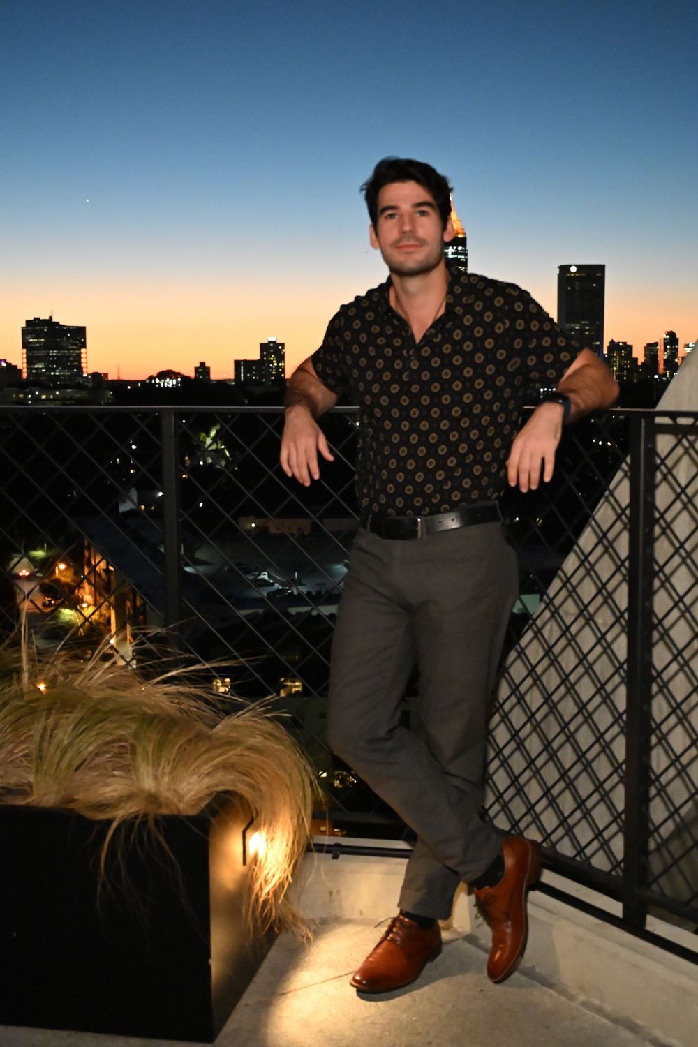 A person sits on a rooftop balcony at sunset with city buildings visible in the background.