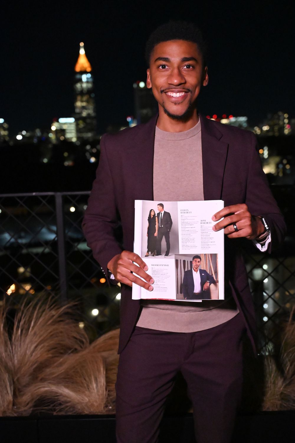 Person in burgundy suit holds up photos against nighttime city skyline backdrop.