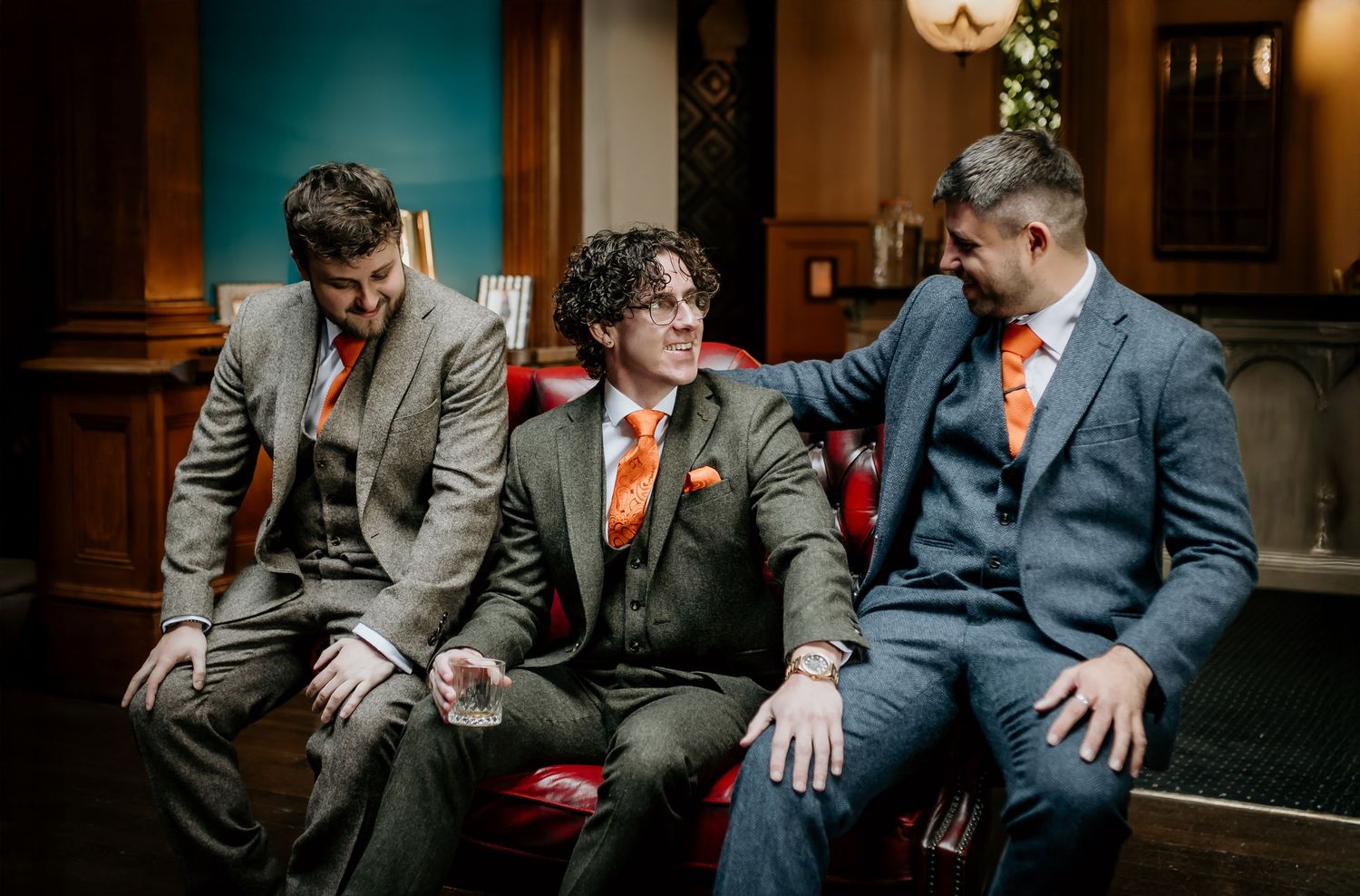 Three groomsmen in suits with orange ties sitting and chatting at wedding venue.