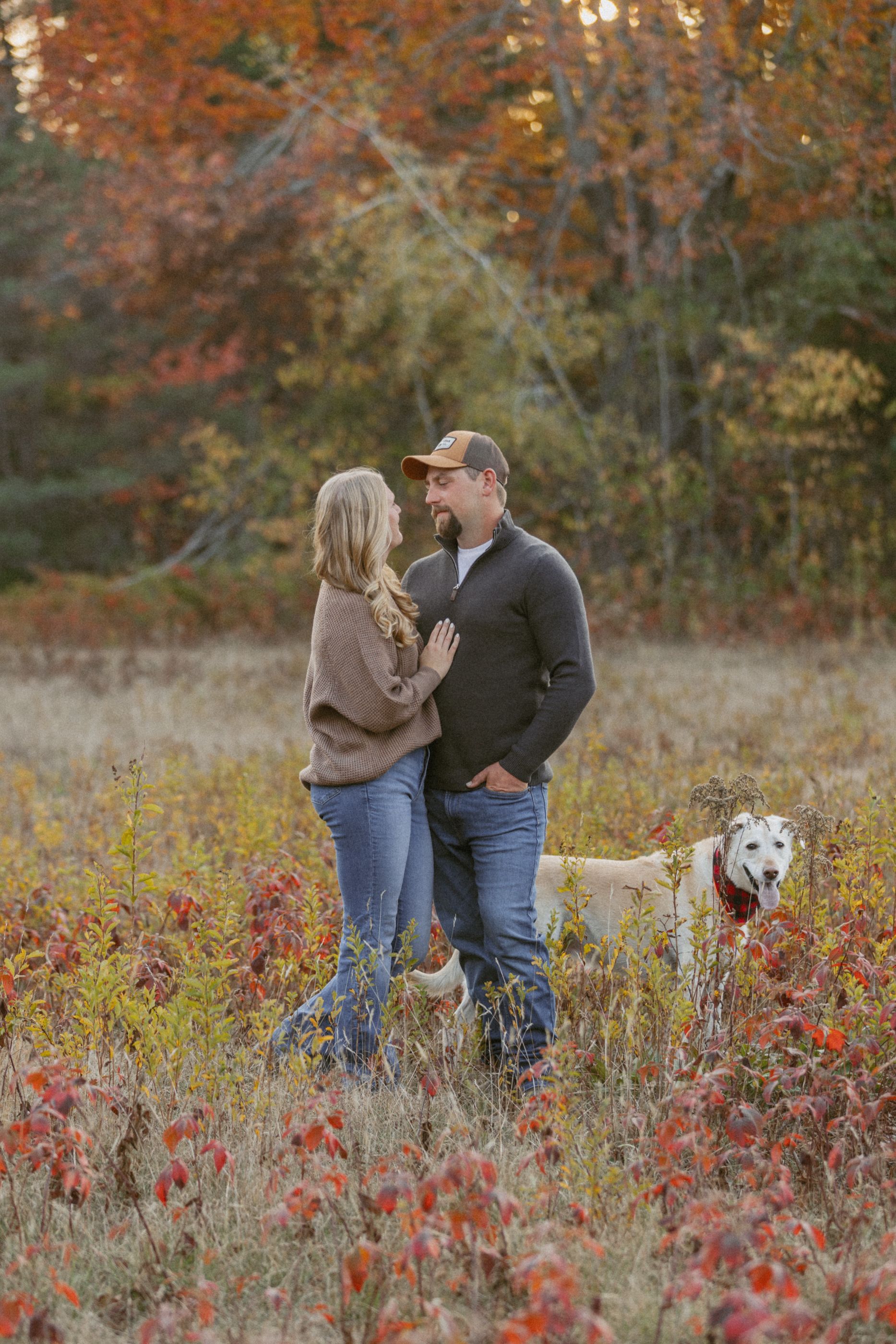 Couple poses in a field of red wildflowers with their white dog during autumn.