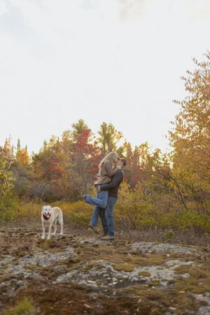 A couple embraces on rocky terrain during autumn with their white dog nearby.