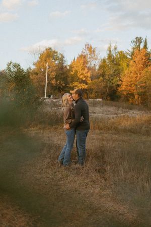 Couple standing together in a field during golden hour with autumn trees in background.