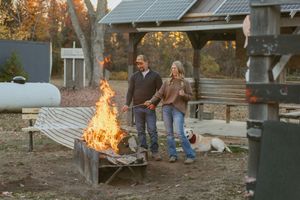 Couple standing by a large bonfire near a covered outdoor structure at dusk.