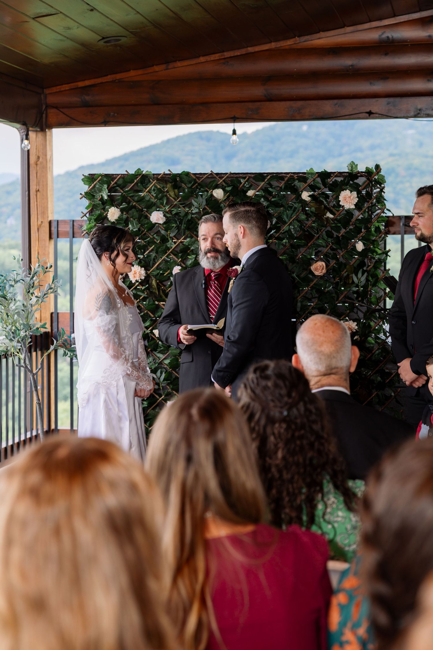 An outdoor wedding ceremony takes place under a wooden pergola with mountain views in the background.