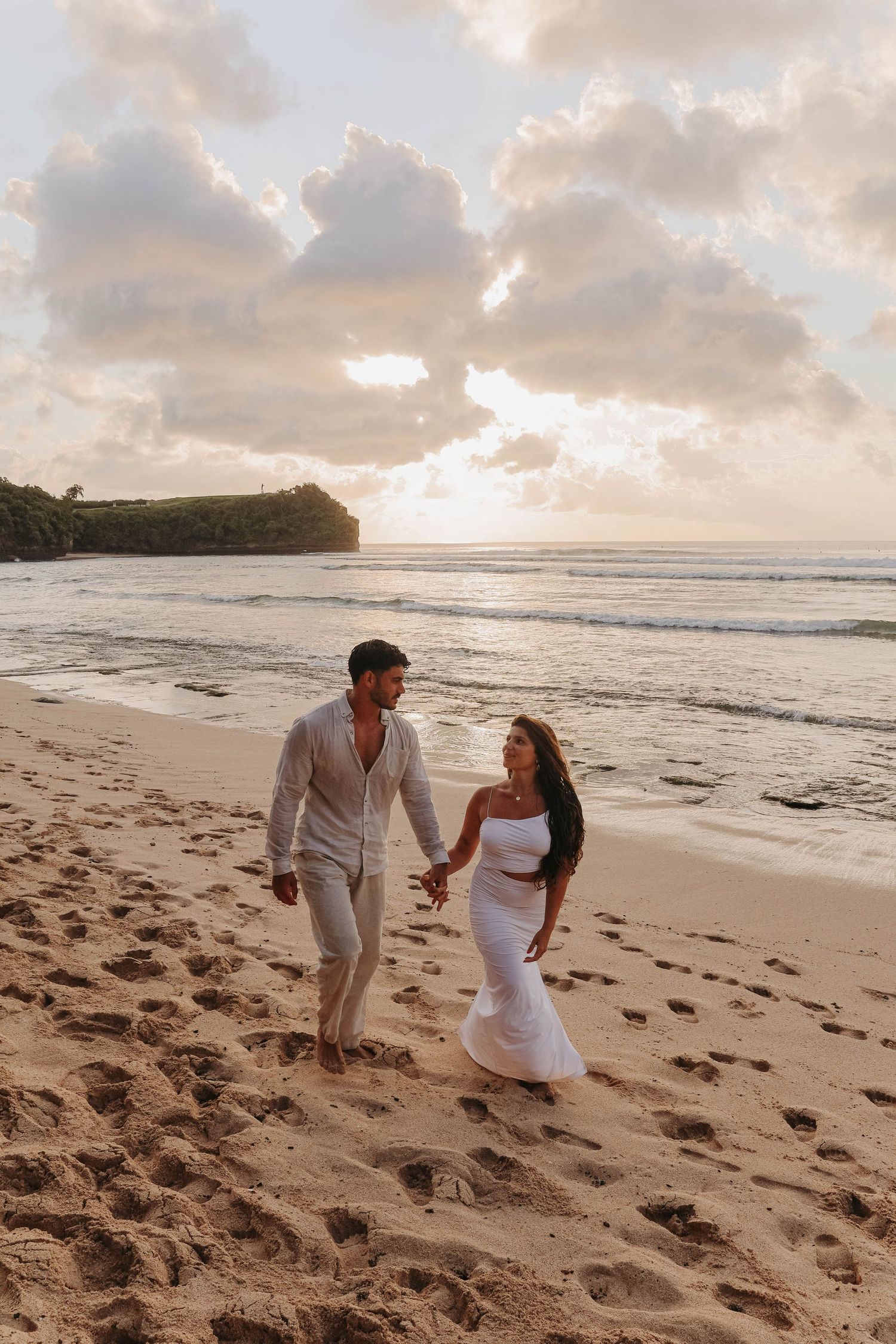 Couple holding hands and walking along sandy beach at sunset with golden clouds and ocean waves.
