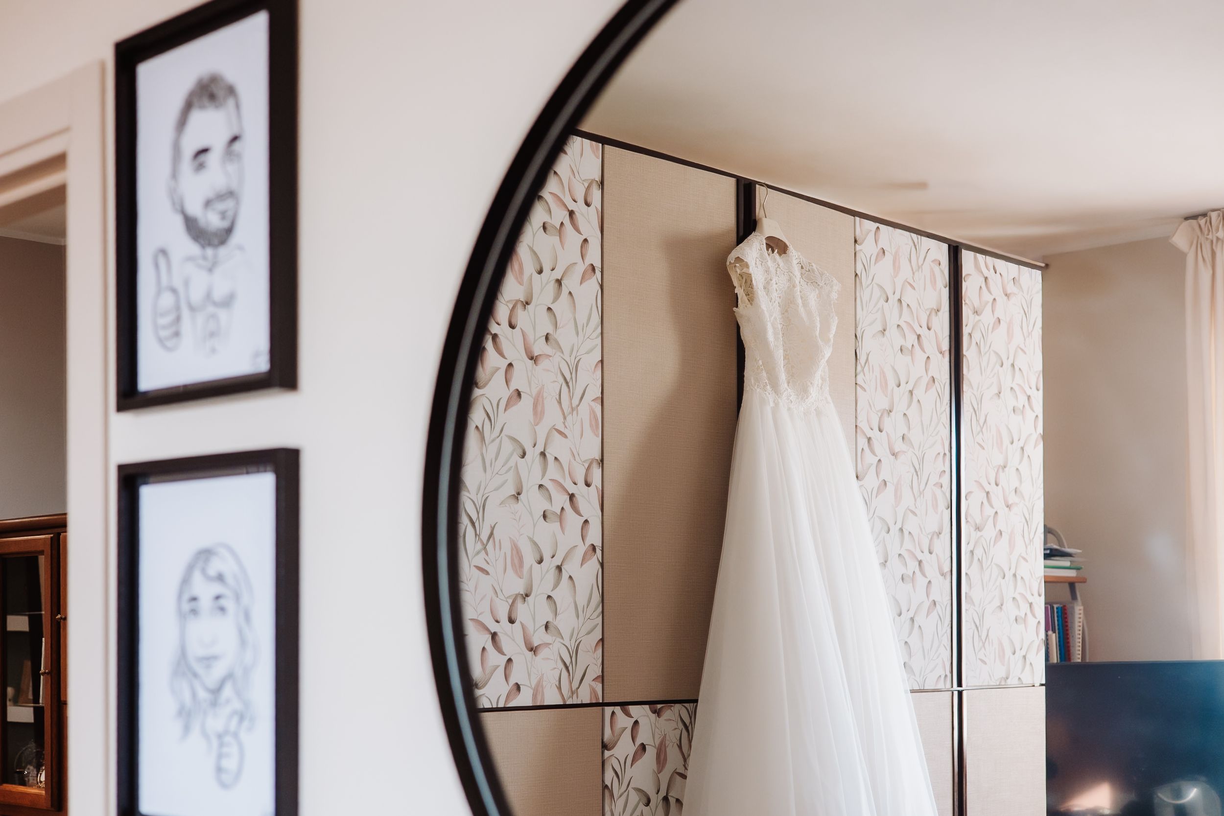A wedding dress hangs elegantly in a bridal suite with a round mirror and floral wallpaper visible in the background.