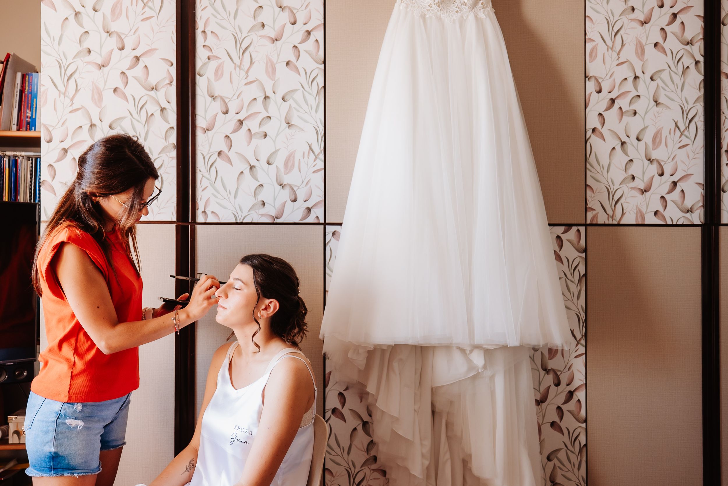 A wedding dress hangs elegantly against a floral wallpapered wall in a getting ready room.