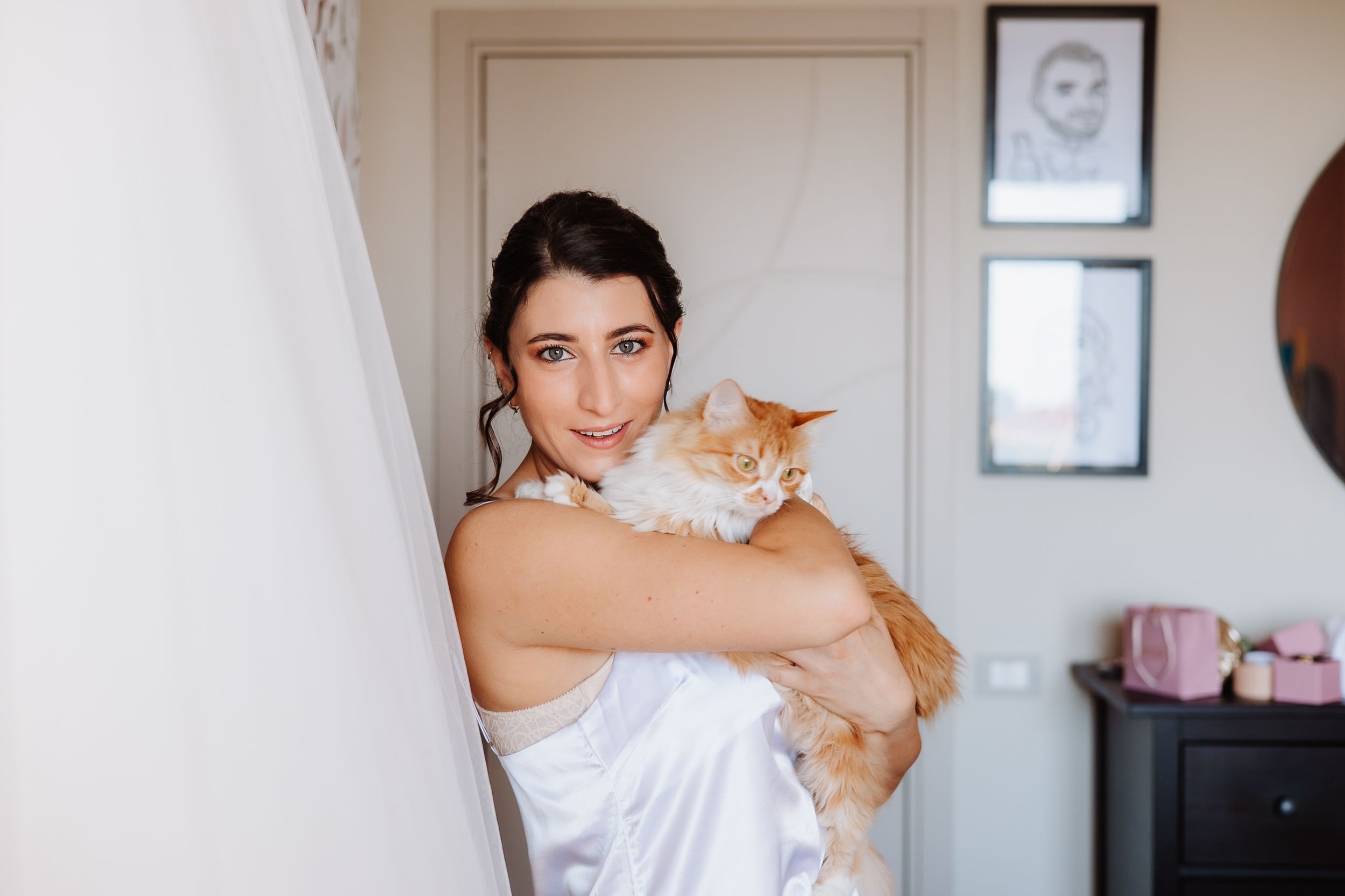 A person in a white dress holds an orange cat while standing in a room with framed photos on the wall.