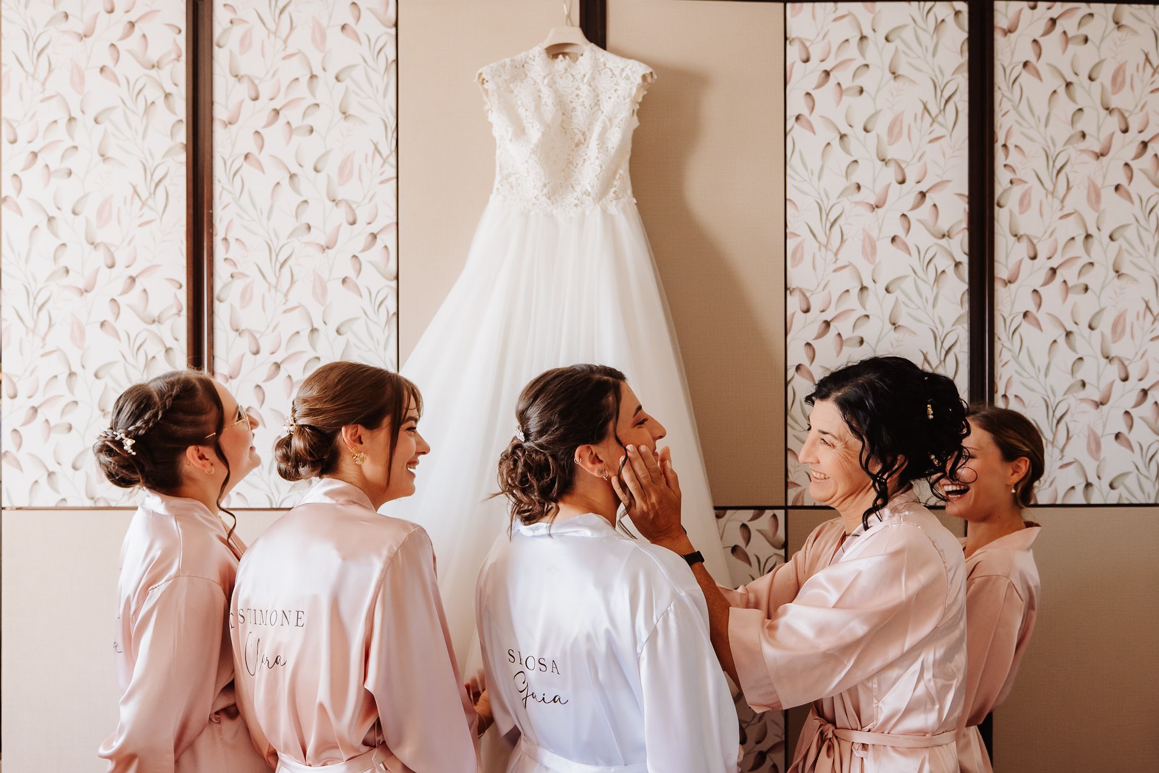 Bridal party in pink robes admiring wedding dress hanging on wall with floral wallpaper.