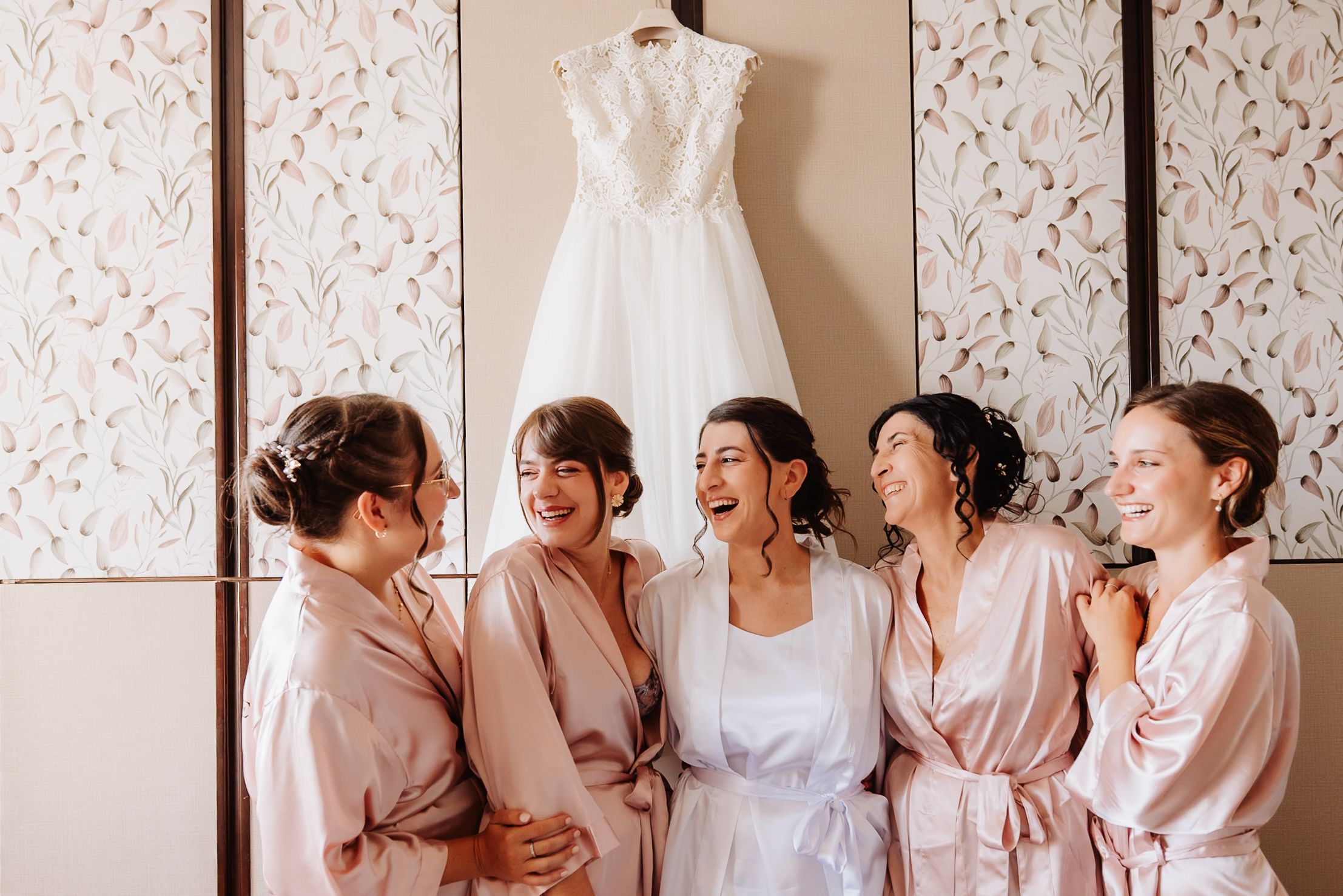 Bridesmaids in matching pink robes gather around a wedding dress displayed on a hanger against floral wallpaper.