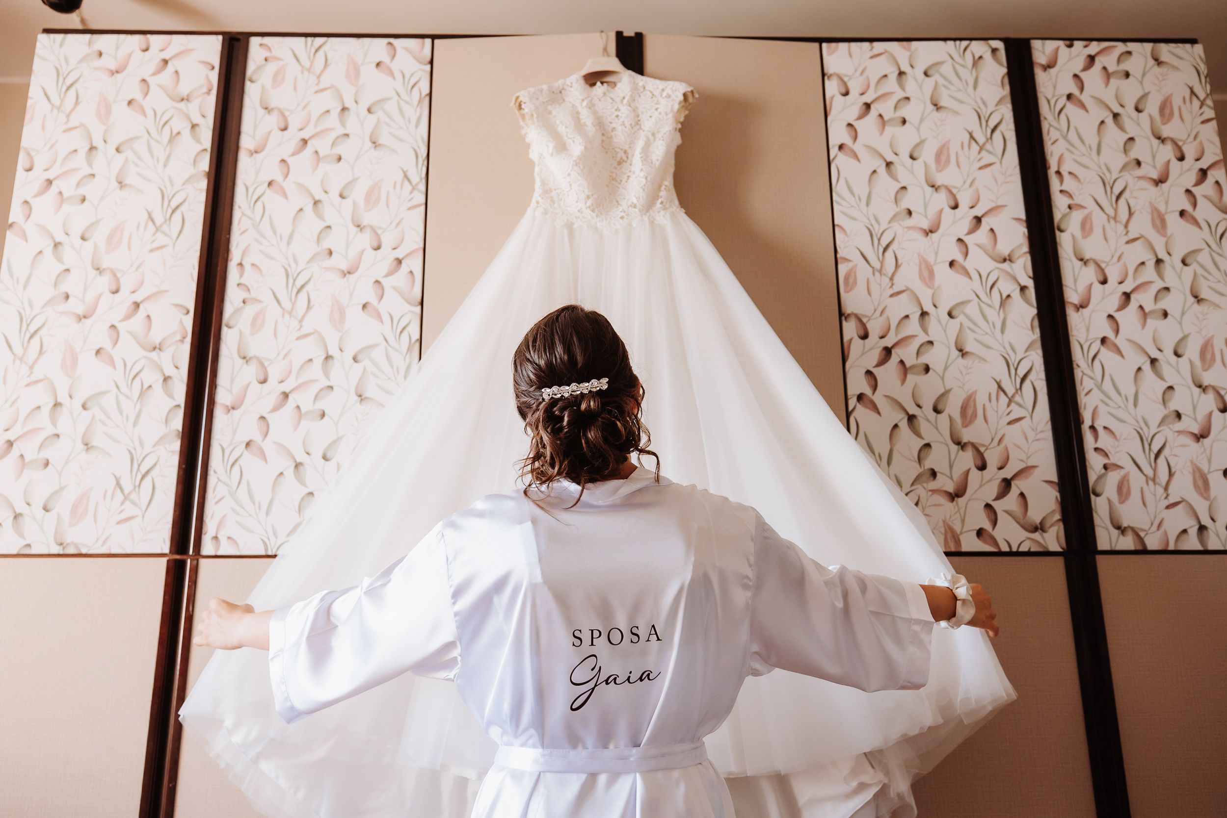 Wedding dress displayed against floral wallpaper while bridesmaids in pink robes gather around it.