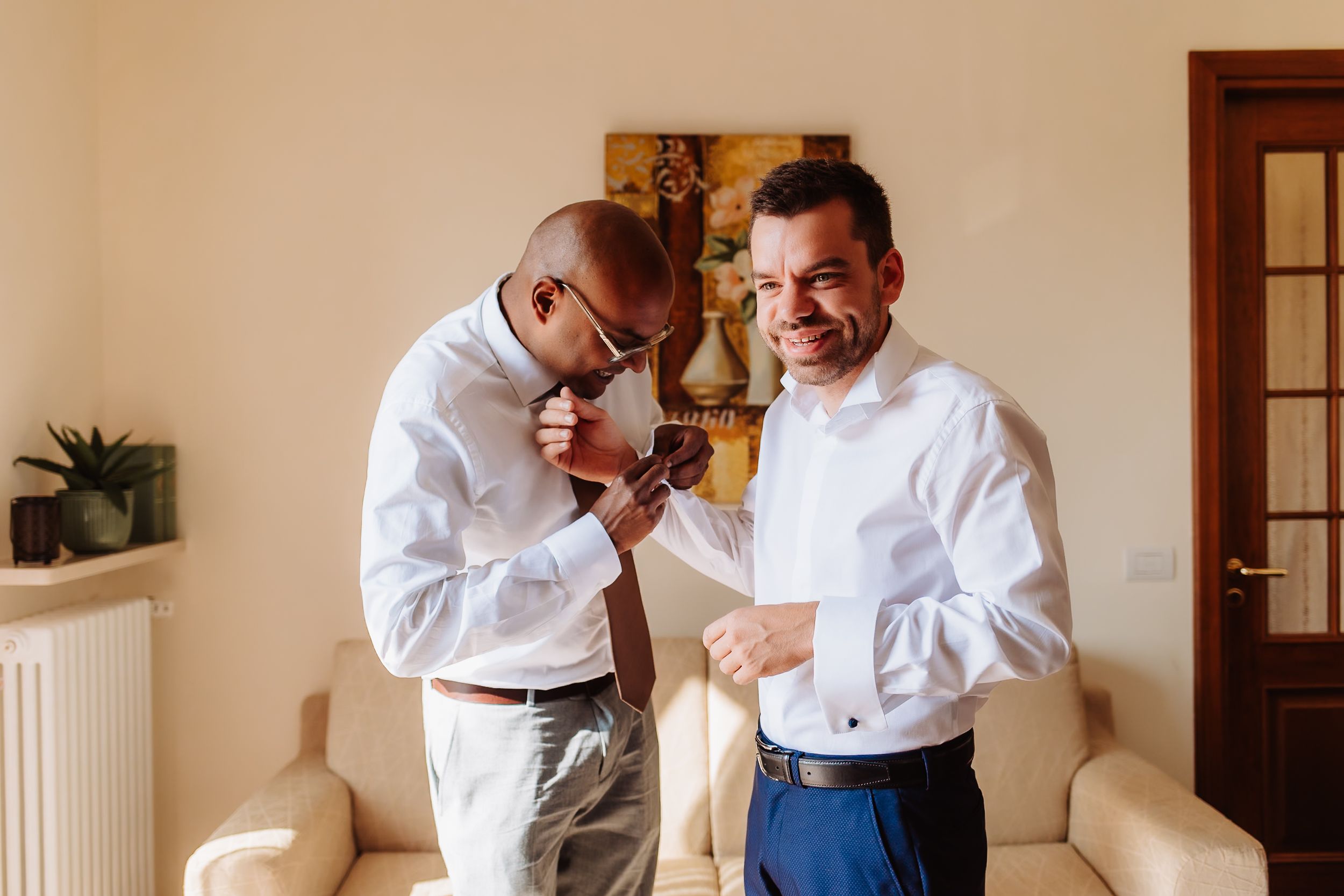 Two people in white shirts stand together while adjusting a tie in a warmly lit interior space.