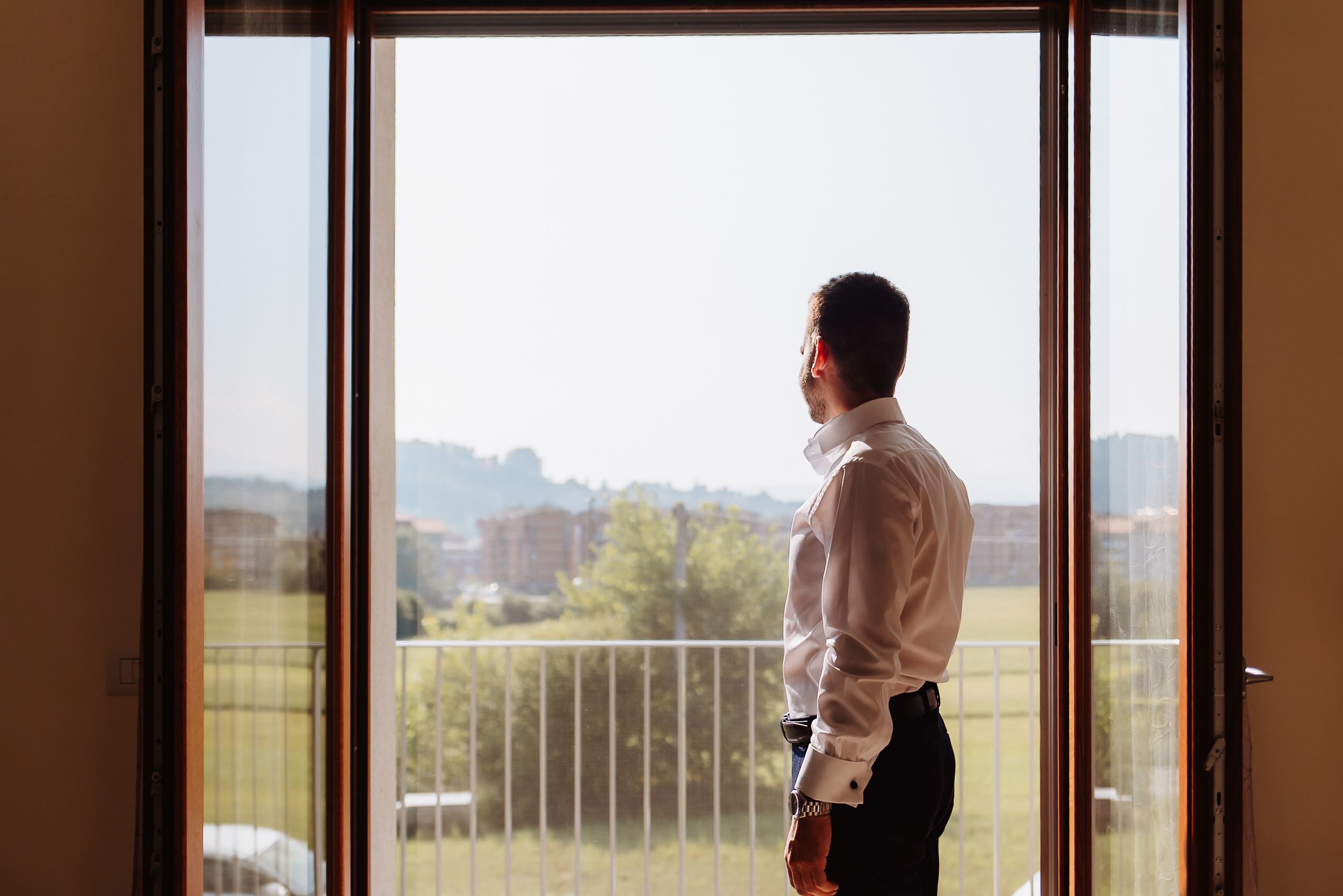 Business person in a white shirt stands at an office window overlooking a city skyline on a sunny day.