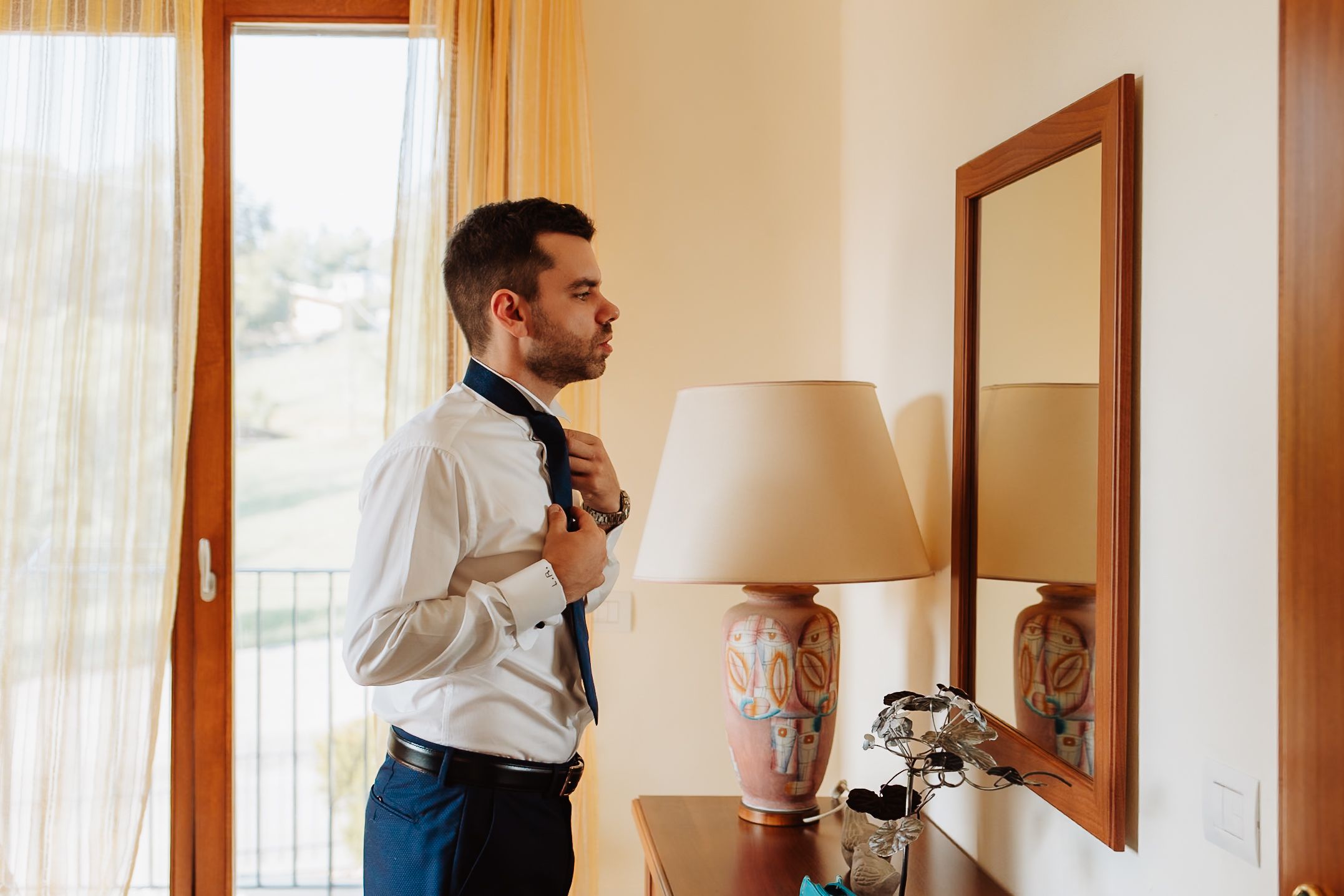 A man in business attire adjusts his tie while looking in a mirror at home.