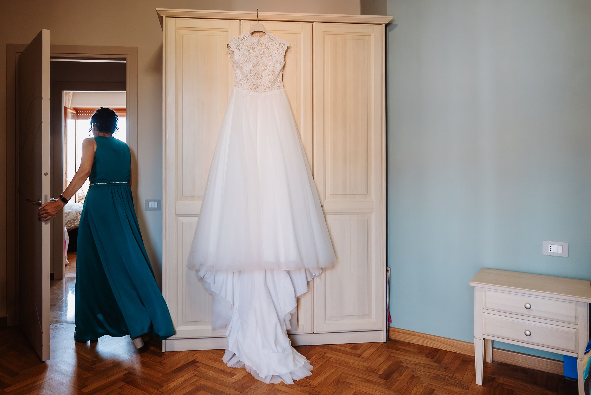 A white wedding dress hangs elegantly against a white wardrobe door in a bridal preparation room.