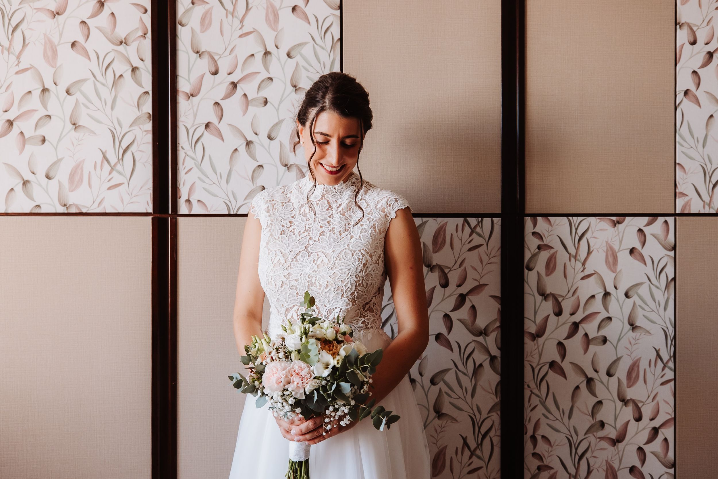 A bride in a sleeveless white lace wedding dress holds a delicate bouquet of pink and white flowers.